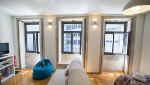 A compact room with a sofa, a bean bag, and a bed. There are two windows providing natural light and a view of buildings outside.