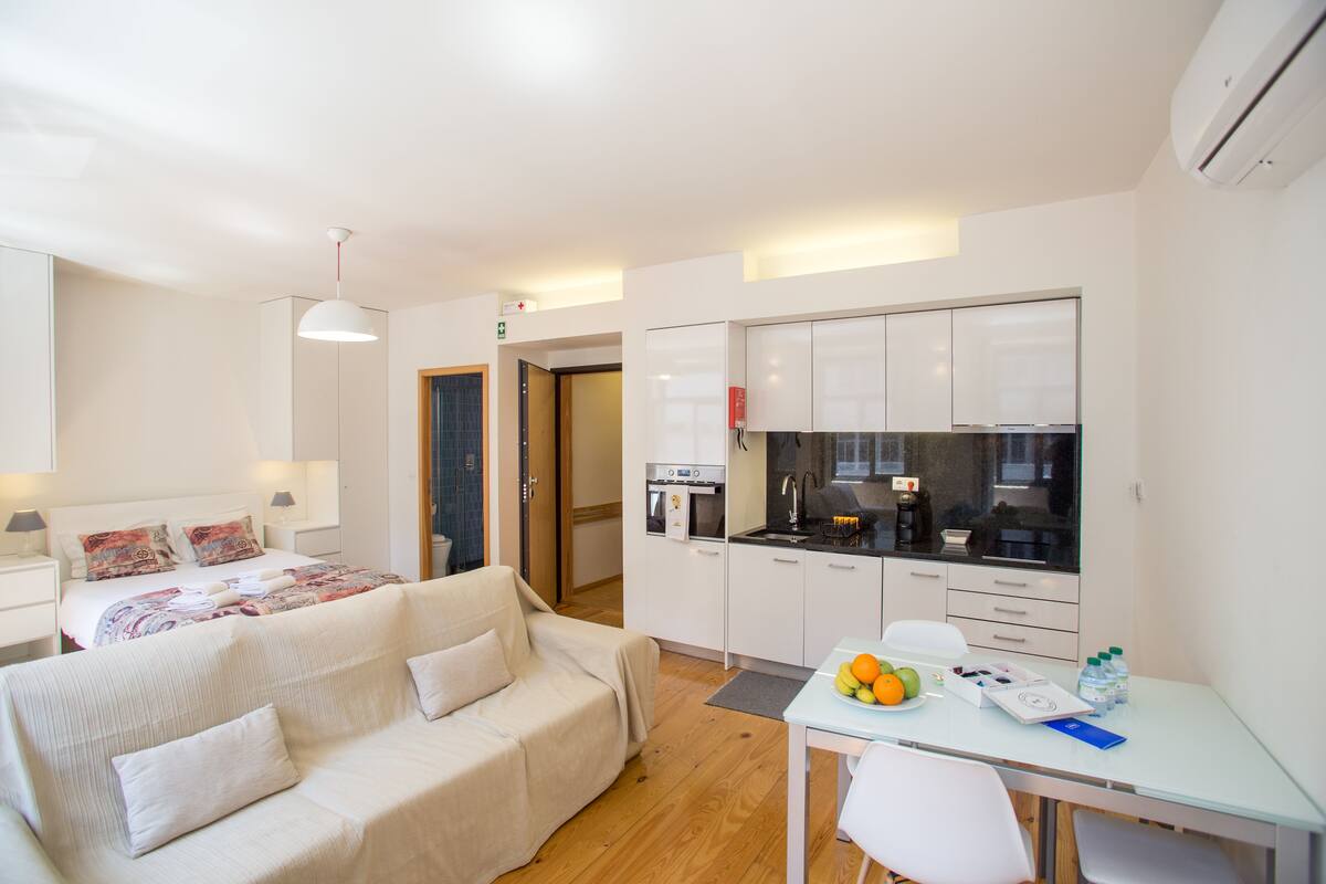 Una habitación de hotel moderna con cama, sofá, zona de comedor con mesa y sillas, kitchenette y baño visible a través de una puerta abierta.