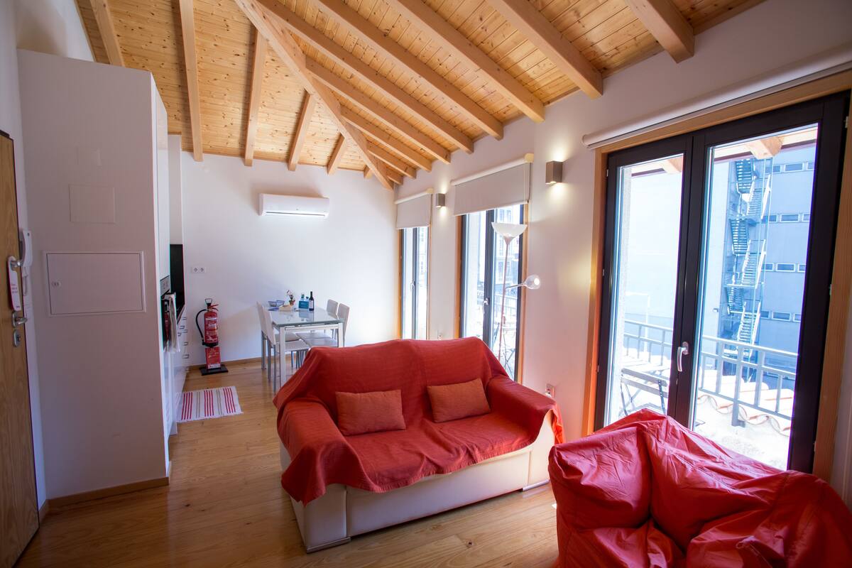 Una habitación amplia con techo de madera, un sofá rojo, una mesa de comedor con sillas y vistas a un edificio a través de grandes ventanales.
