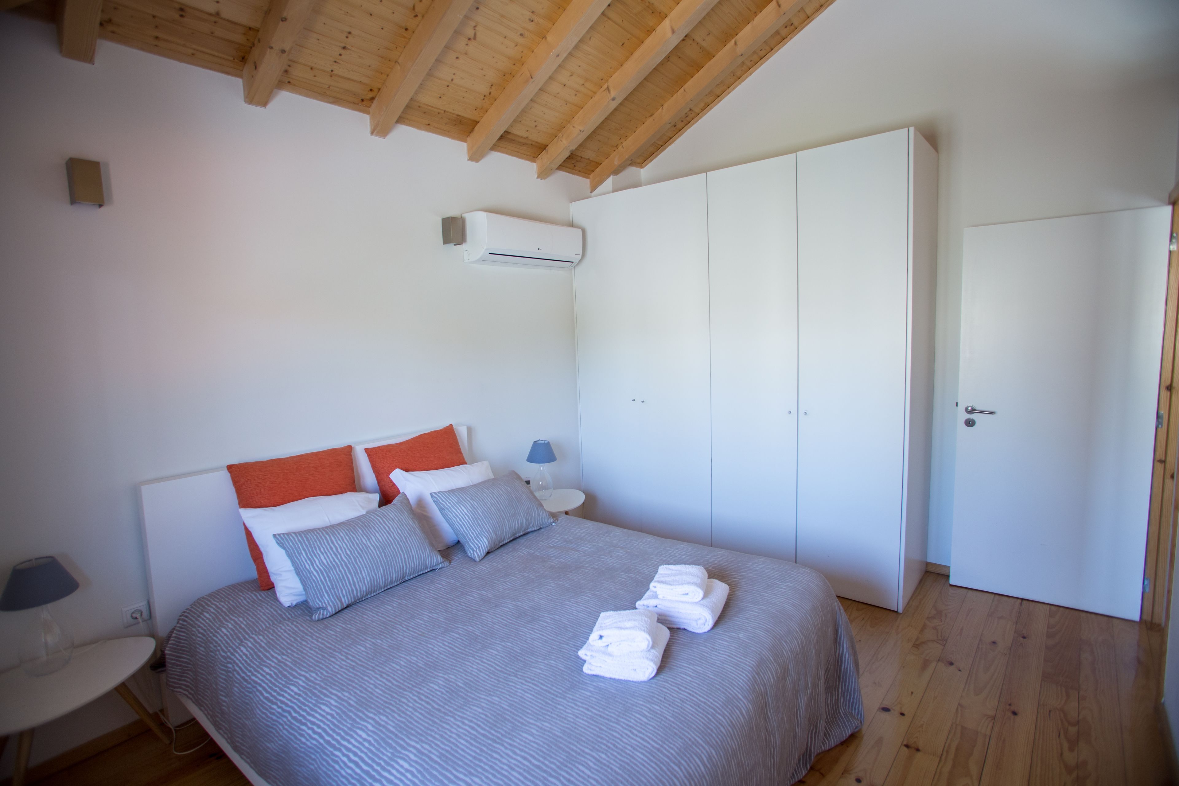A bedroom with a bed, pillows, a bedside table, a lamp, a wardrobe, and an air conditioning unit.