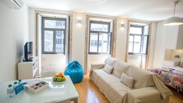 A well-lit room with a white sofa, a small table with a fruit bowl, a blue bean bag, and a TV on a white cabinet.