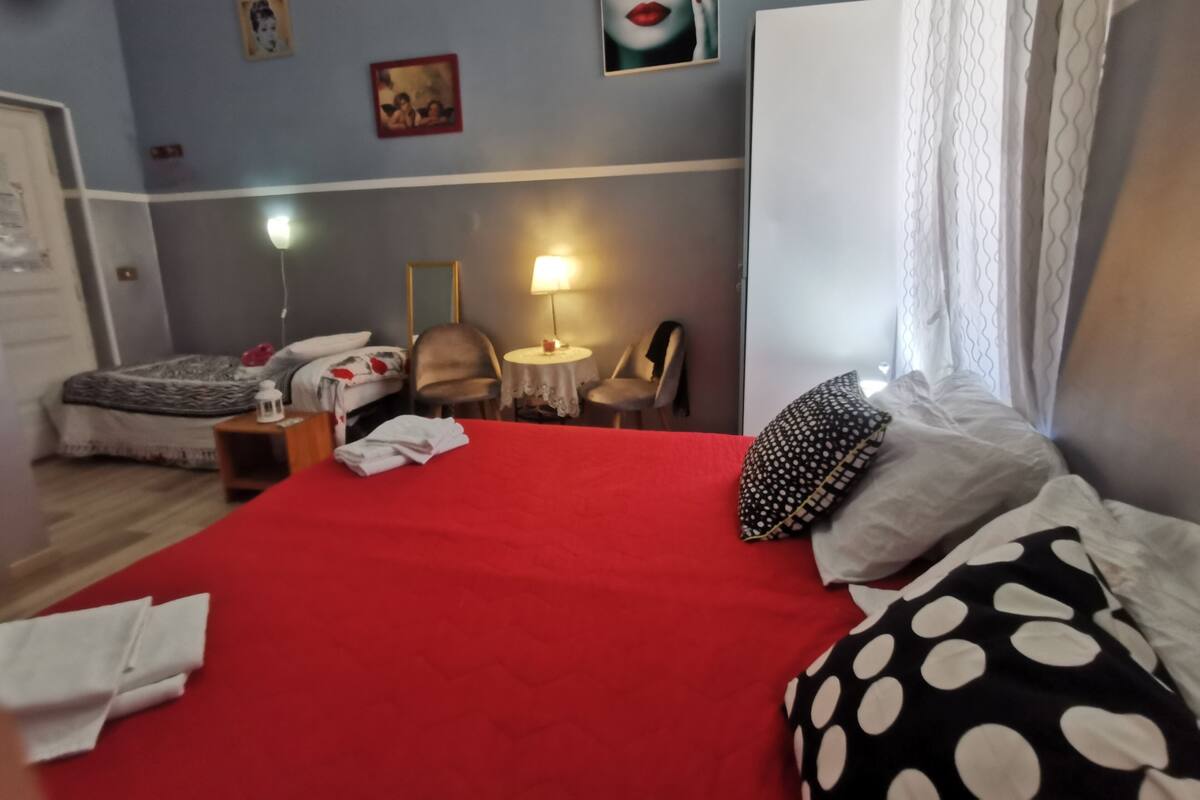 A hotel room with a red bed, a chair, a small table with a lamp, and a painting on the wall.