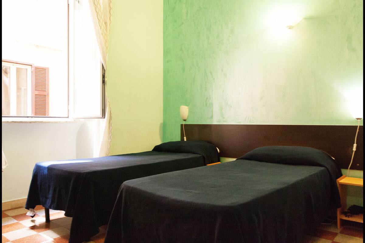 A room with two single beds, a window with curtains, a wall lamp, and a checkered floor.
