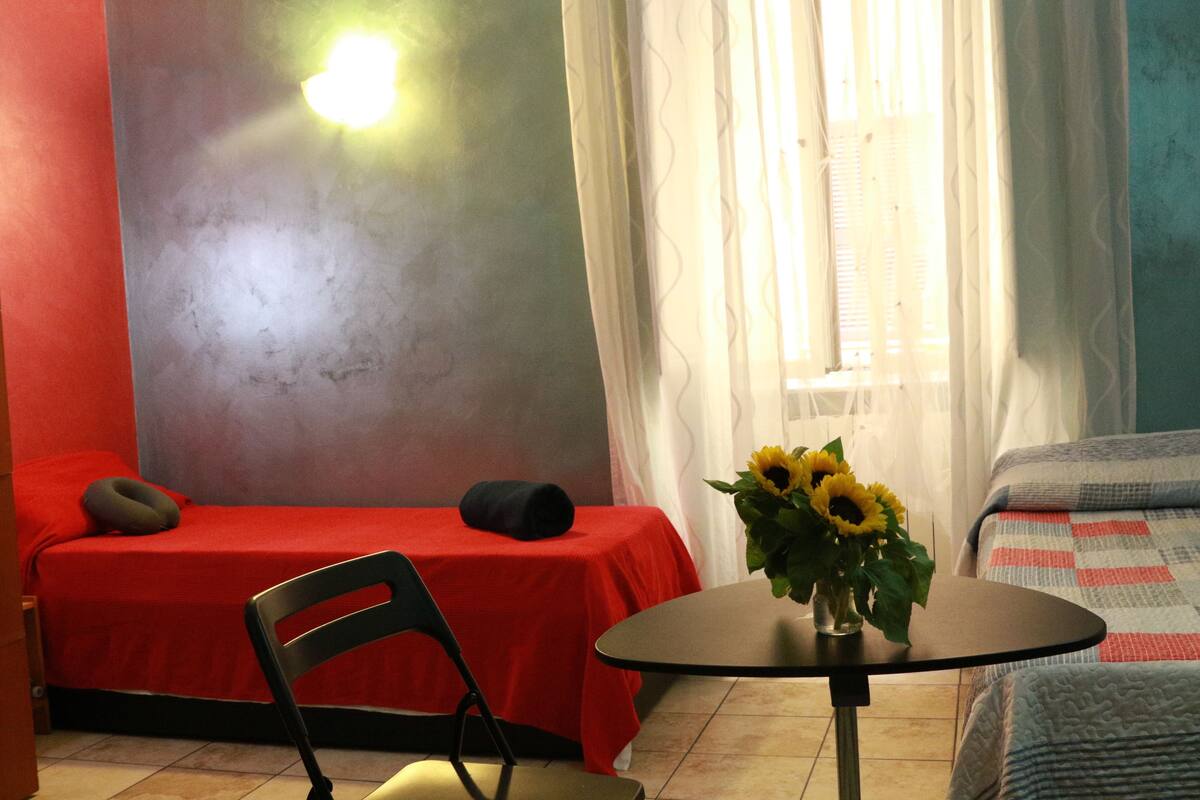 A room with a single bed, a chair, a small table with a vase of sunflowers, and a window with curtains.