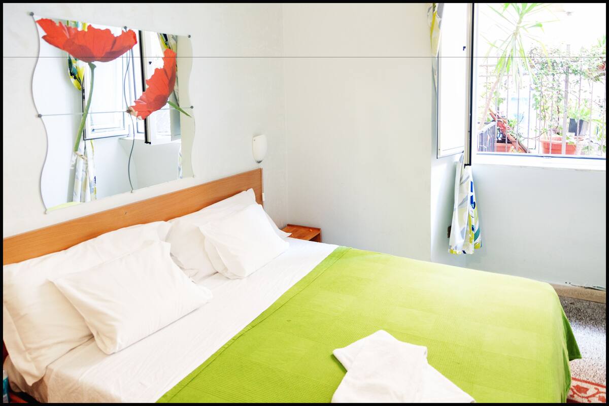 A hotel room with a bed, a green bedspread, white pillows, a wooden headboard, a nightstand, a mirror, and a window with a view of plants outside.