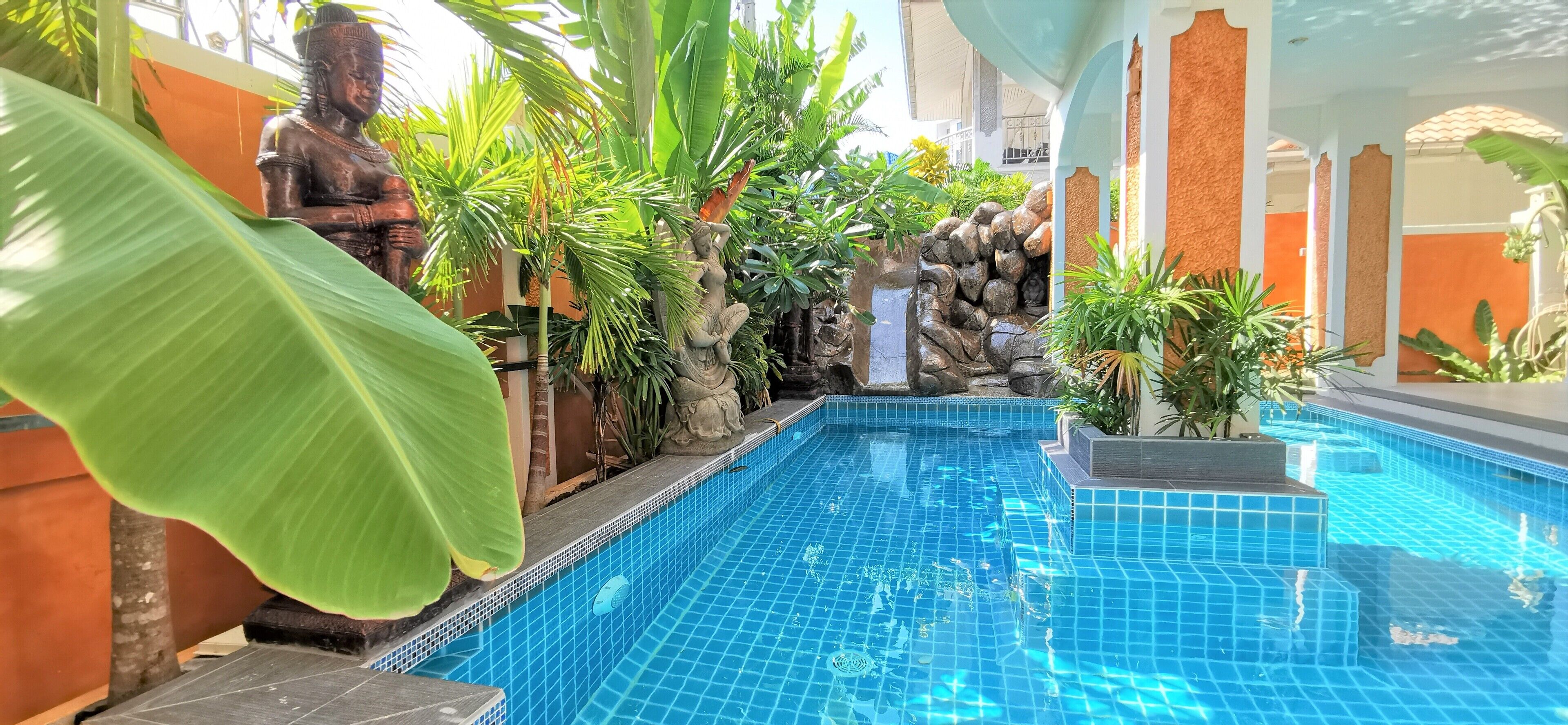 Ein Schwimmbecken mit blau gefliesten Rändern, umgeben von tropischer Bepflanzung und einer Statue.