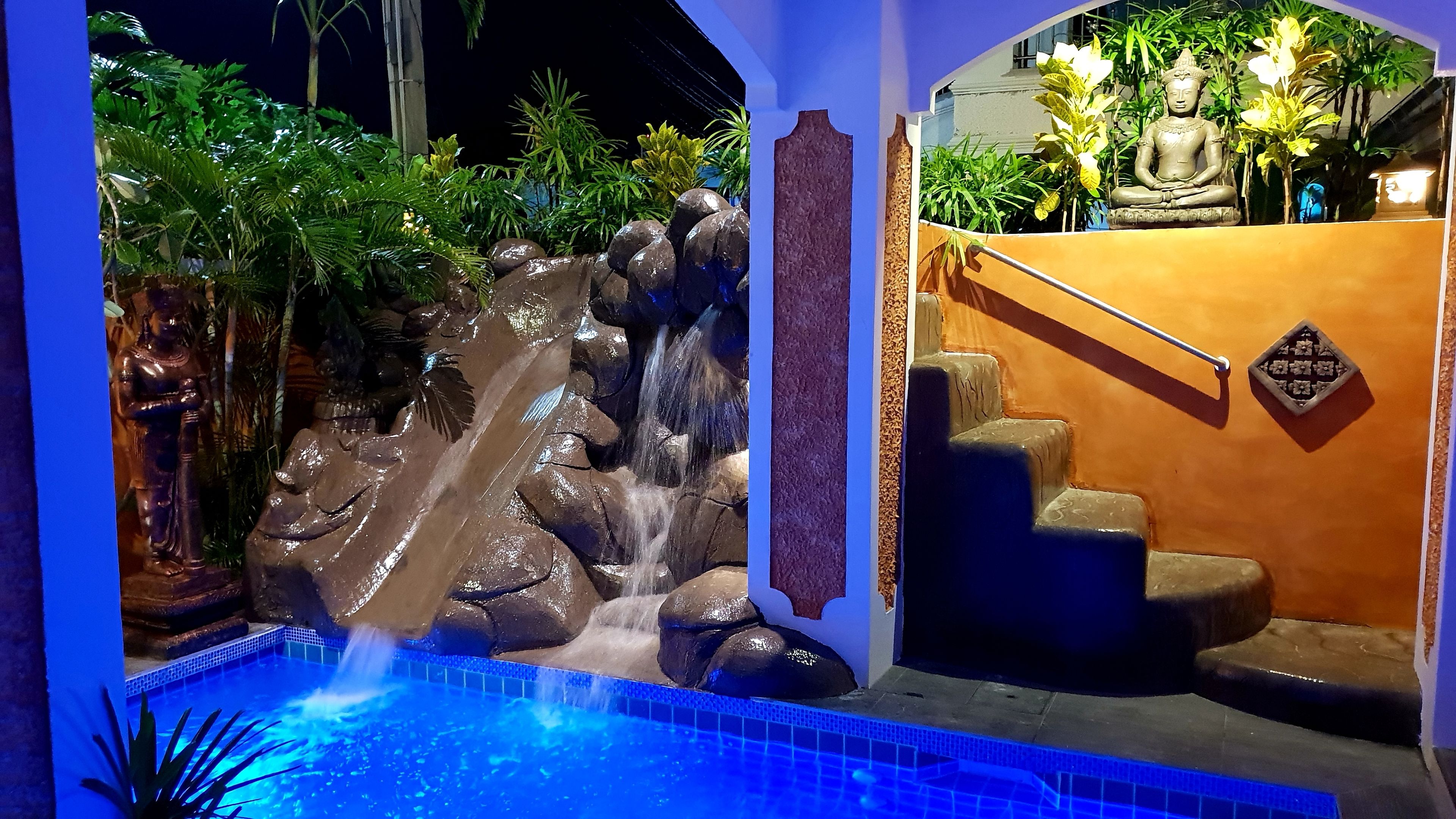 Una fuente con cascada y estatua, rodeada de abundante vegetación y una escalera que conduce a un nivel superior.