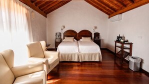 A bedroom with a wooden ceiling, two beds, a chair, a nightstand, a lamp, and a trash bin. - Hotel Rural Casa de los Camellos (Aguimes)