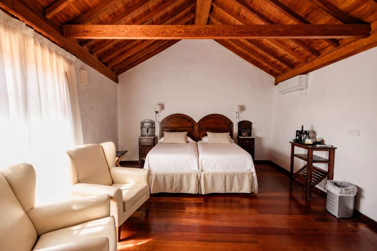 A bedroom with a wooden ceiling, two beds, a chair, a nightstand, a lamp, and a trash bin.