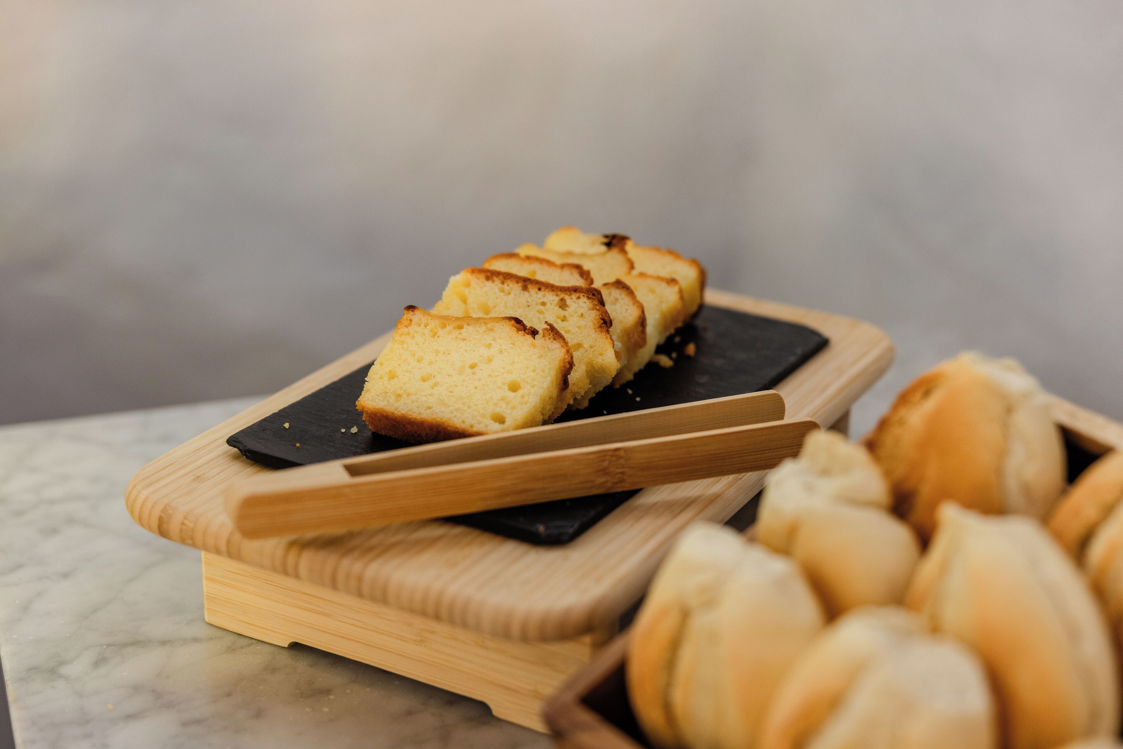 Een brood op een houten snijplank met een mes, en een mand met broodjes op de achtergrond.