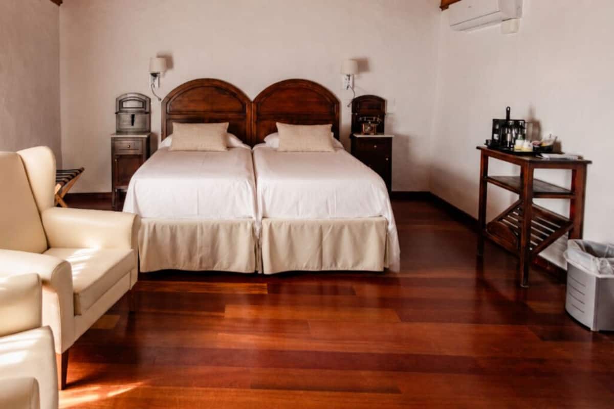 A double bed with wooden headboards, a chair, a small table, and a trash bin in a hotel room.