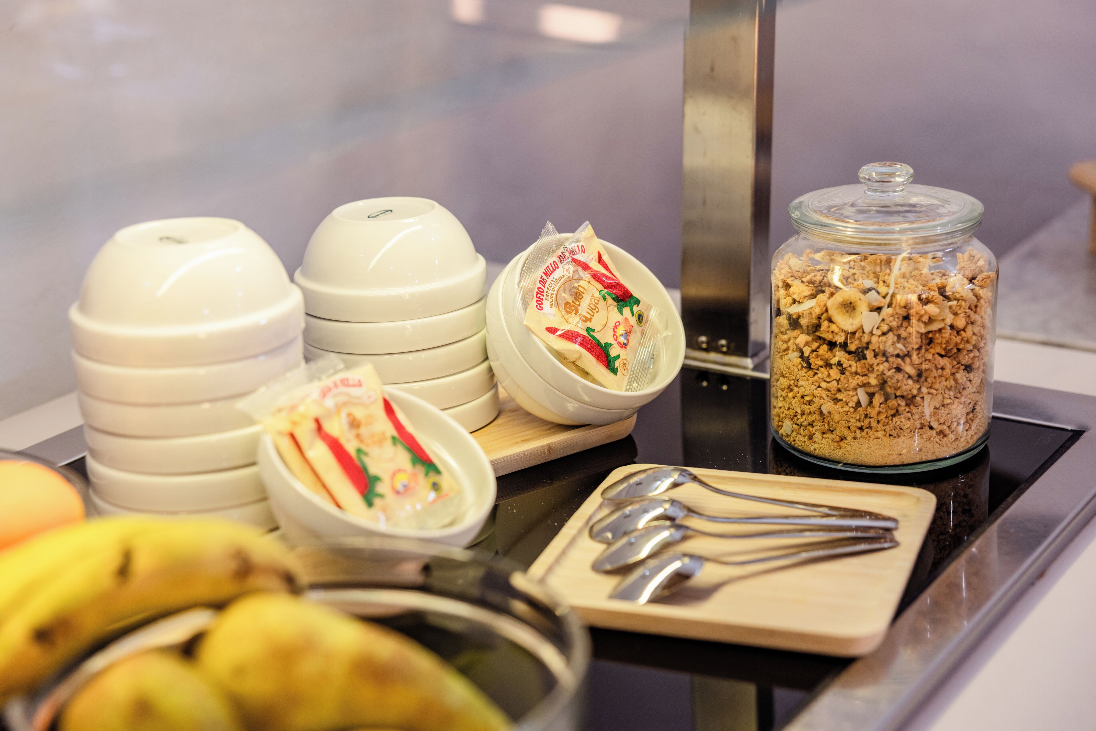 Un bufé de desayuno con tazones de comida, un tarro de granola y utensilios sobre una encimera.