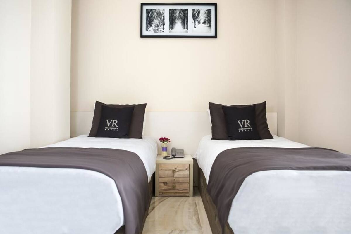 A hotel room with two beds, each with a pillow labeled 'VR'. There is a small bedside table with a vase of flowers and a phone.