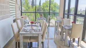 A dining area with a large table set for six, surrounded by chairs, with a view of trees and buildings through large windows. - Villa Residence Hotel (Tbilisi)