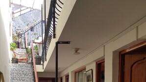 Un estrecho pasillo con suelo de baldosas, una puerta de madera y una escalera con barandillas de metal que sube a un nivel superior.