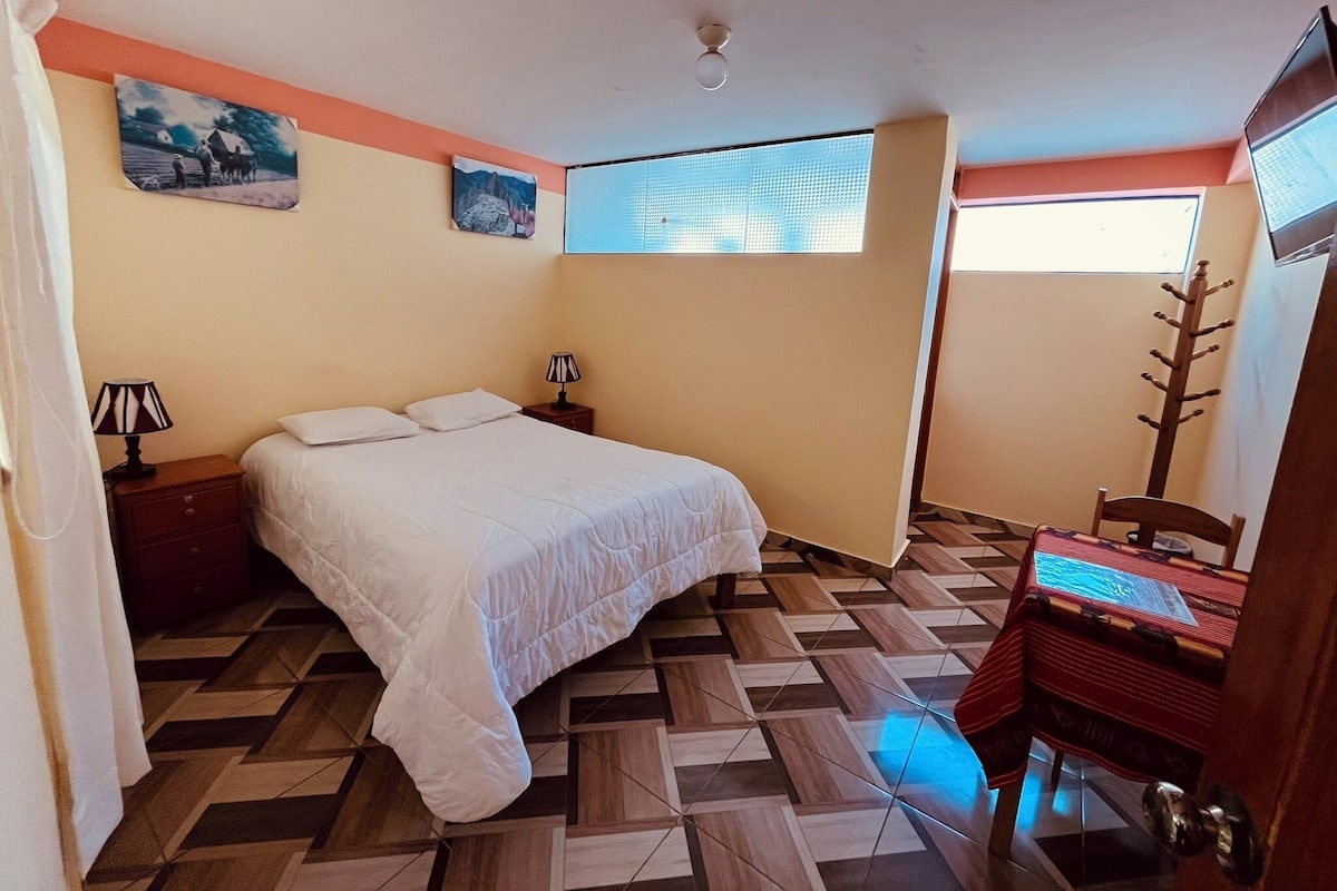 Ein Schlafzimmer mit einem Bett, zwei Nachttischen, einem Schreibtisch mit Stuhl, einem Fenster und zwei Wandbildern.
