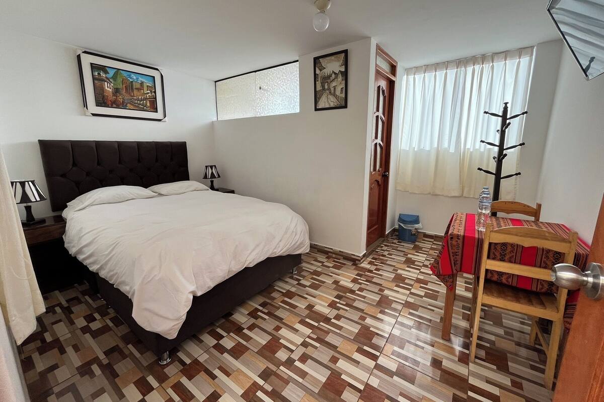 Ein Schlafzimmer mit Bett, Nachttisch, Lampe, Stuhl, Tisch, einer Wasserflasche, einer Tür und einem Fenster mit Vorhängen.