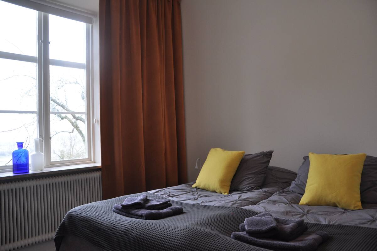 A hotel room with a large window, a radiator, a bed with grey and yellow pillows, and a grey blanket.