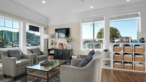 A spacious living area with two grey armchairs, a glass coffee table, a black cabinet, and a bookshelf with wicker baskets. - Looking Glass Inn (Lincoln City)