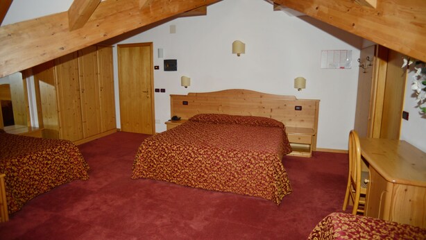 A room with a sloped ceiling, wooden beams, a bed with a red bedspread, a wooden desk, and a chair. - Hotel Canada (Andalo)