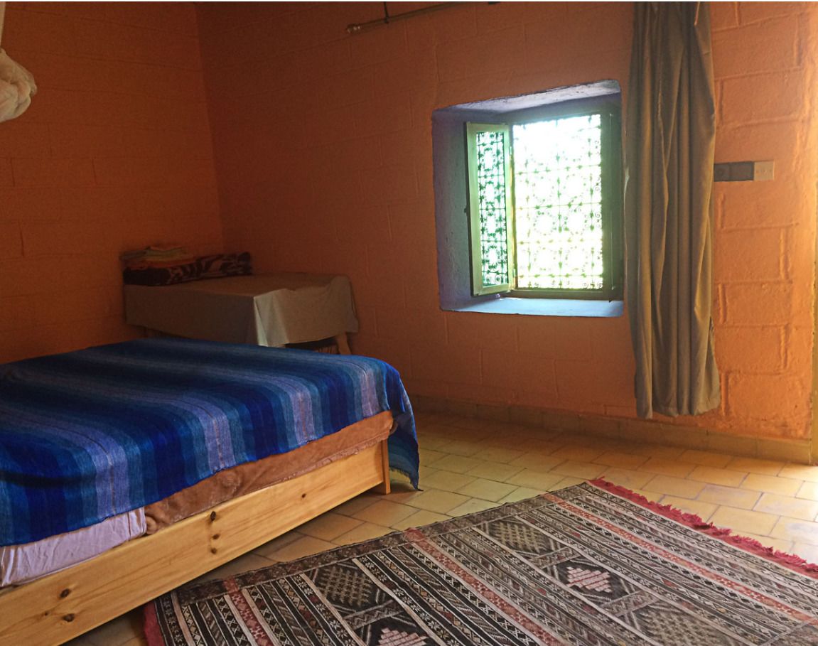 A bedroom with a bed, a small table, a window with curtains, and a patterned rug.