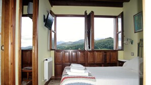 Une chambre avec un grand lit, une vue sur la campagne par de grandes fenêtres ouvertes et des lambris en bois sur les murs.