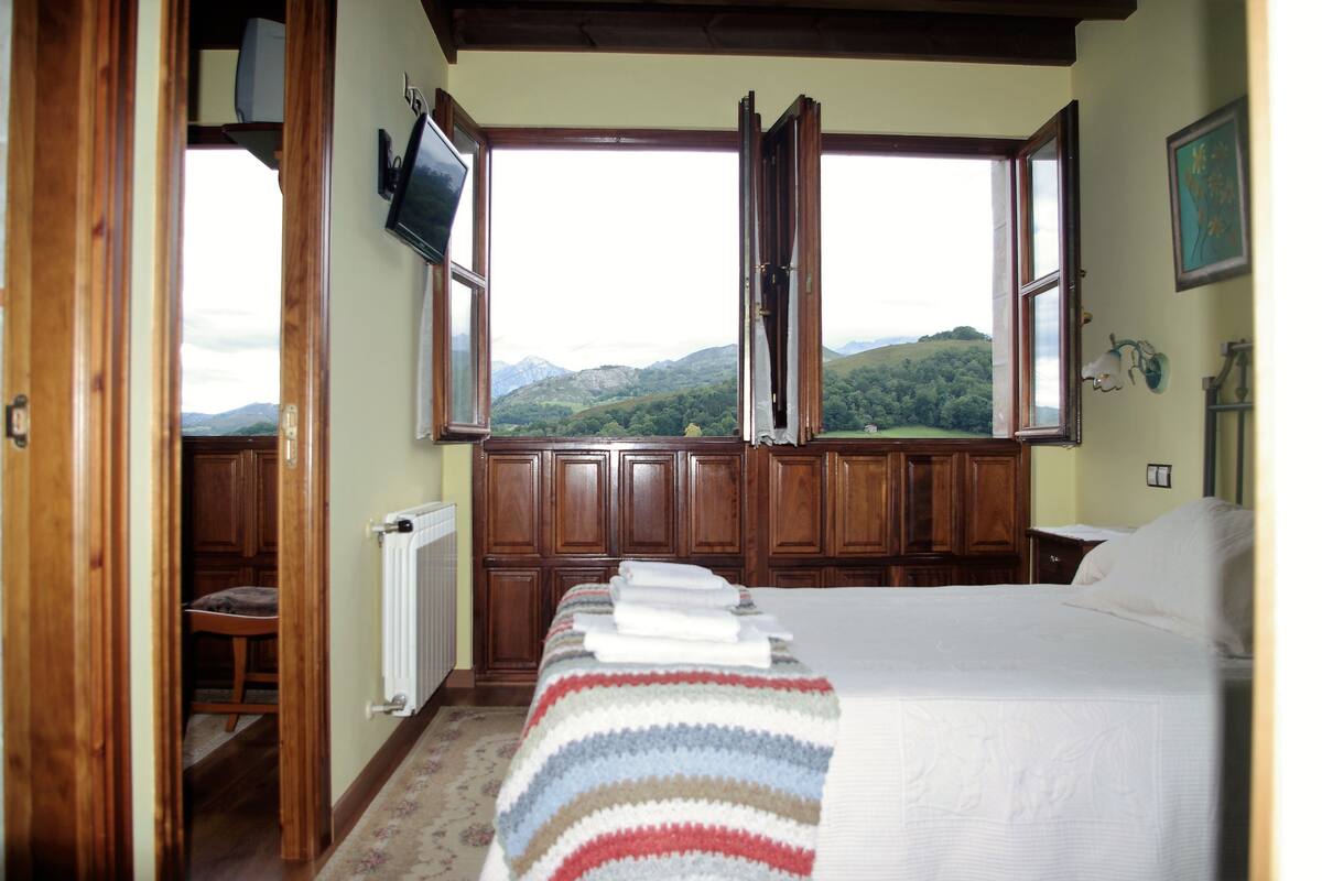 Une chambre avec un grand lit, une vue sur la campagne par de grandes fenêtres ouvertes et des lambris en bois sur les murs.