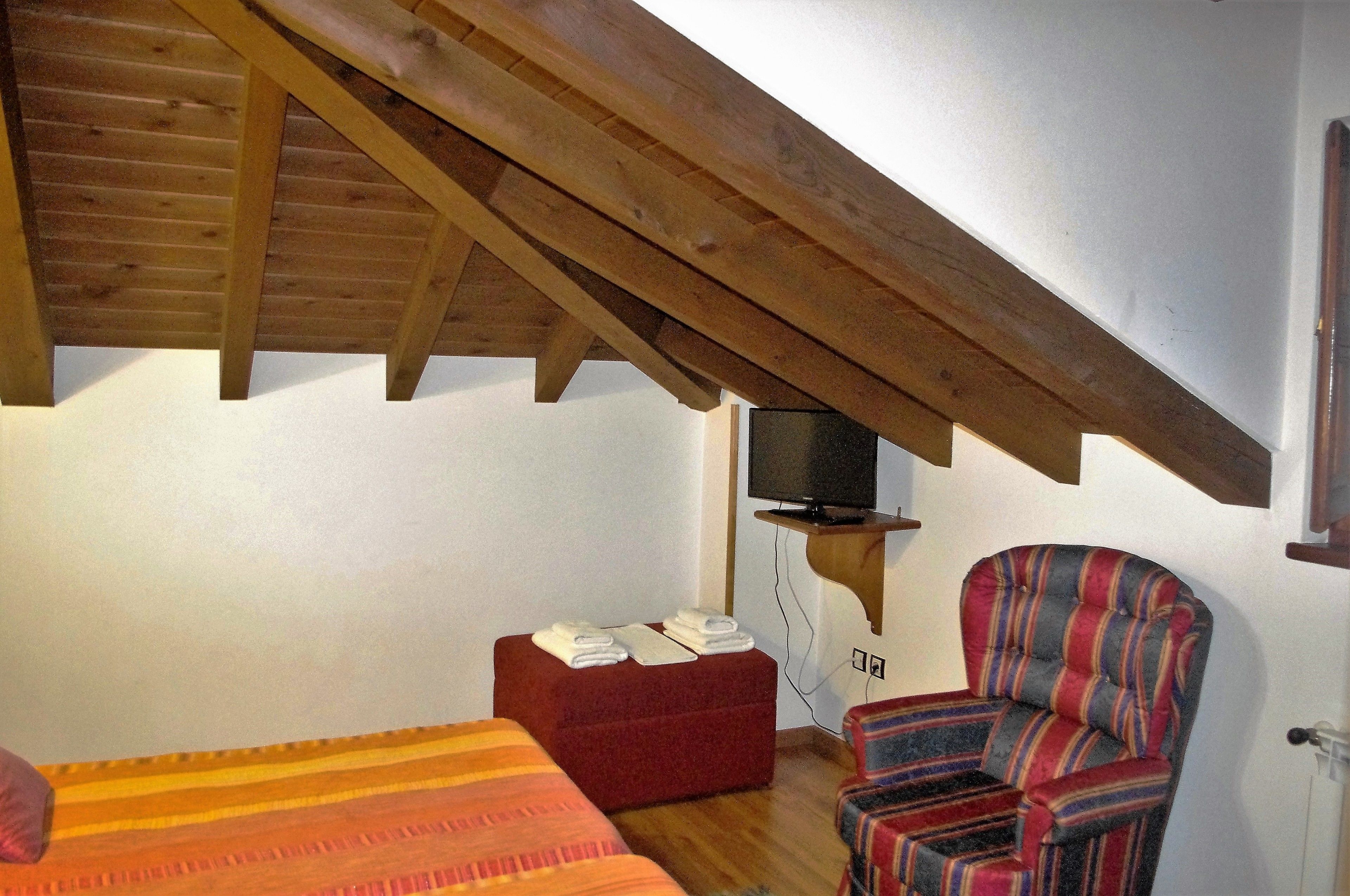 Une pièce avec un plafond en pente, un lit, un pouf rouge, un fauteuil à rayures et un téléviseur posé sur un meuble.