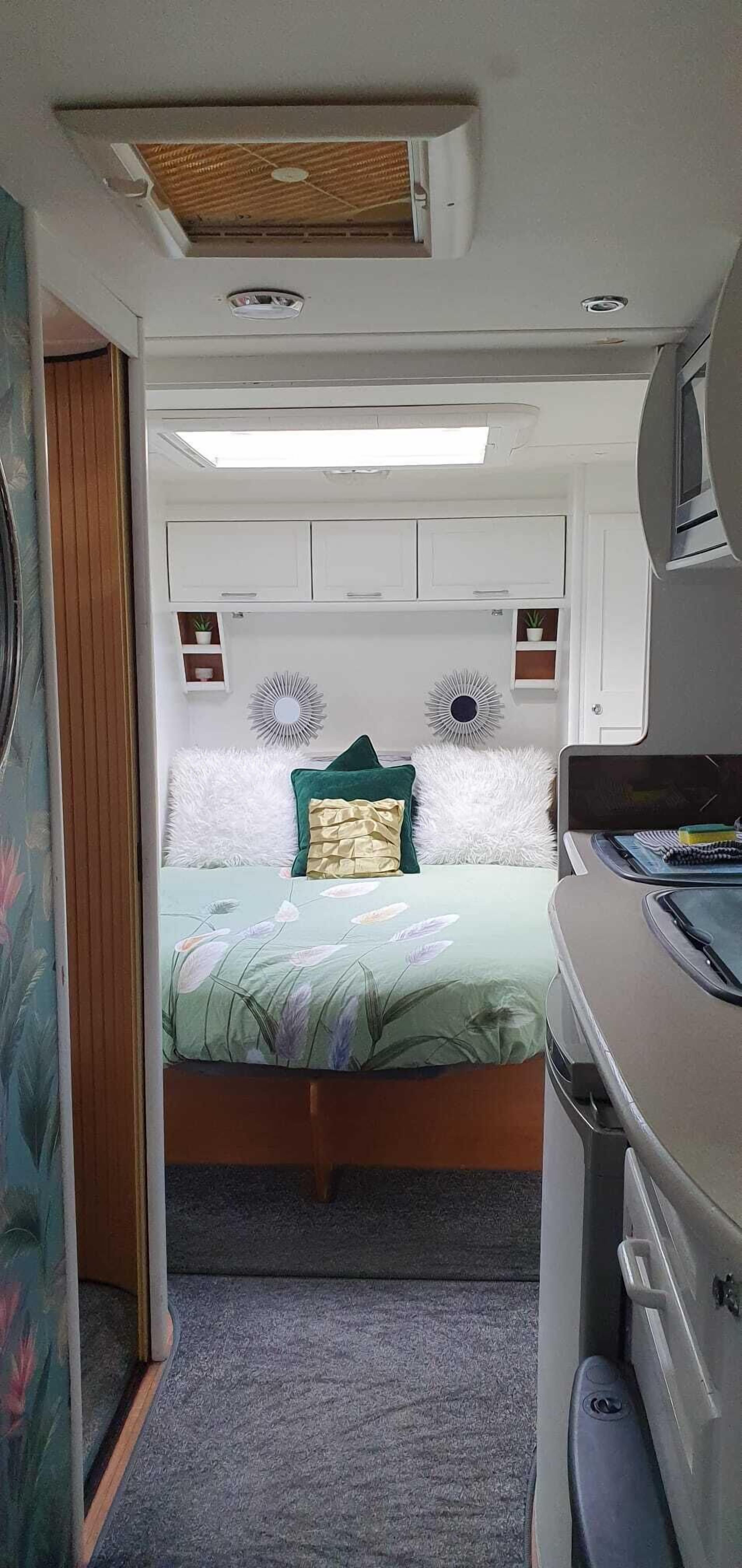 Une chambre compacte dans un camping-car, comprenant un lit, une petite étagère avec des plantes et un ventilateur de plafond.
