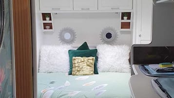Une chambre compacte dans un camping-car, comprenant un lit, une petite étagère avec des plantes et un ventilateur de plafond.