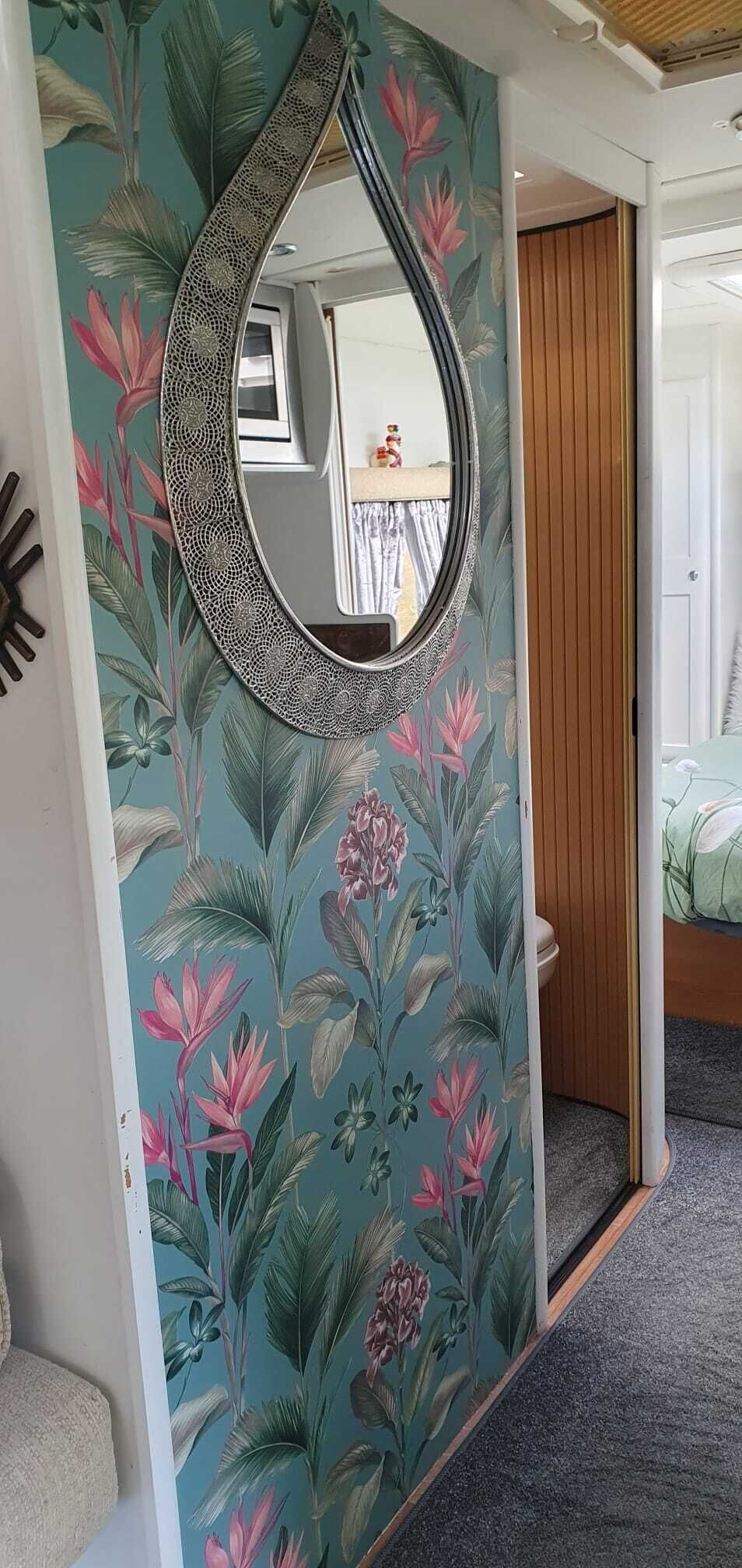 A room with a floral wallpaper, a decorative mirror, and a bed with a green bedspread.