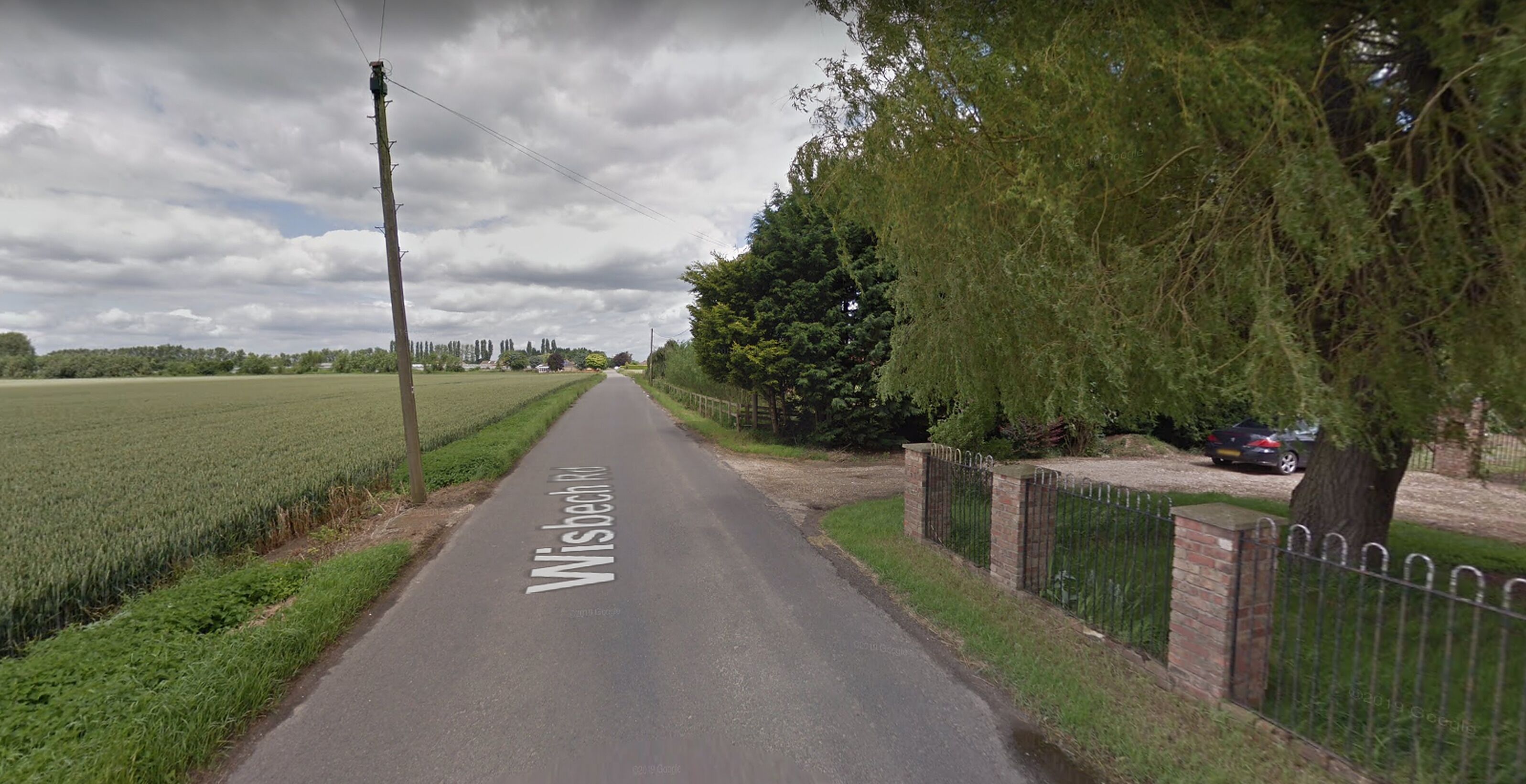 Une route de campagne appelée « Wisbech Rd », bordée de champs de chaque côté et d’une clôture sur la droite.