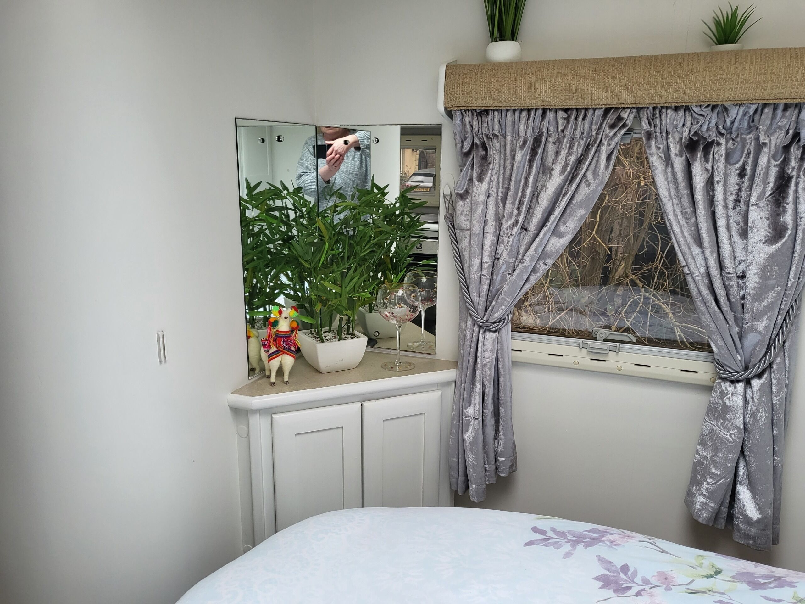 Ein Schlafzimmer mit einem Bett, einem Fenster mit grauen Vorhängen, einem Spiegel, einer Topfpflanze und einem dekorativen Gegenstand auf der Fensterbank.