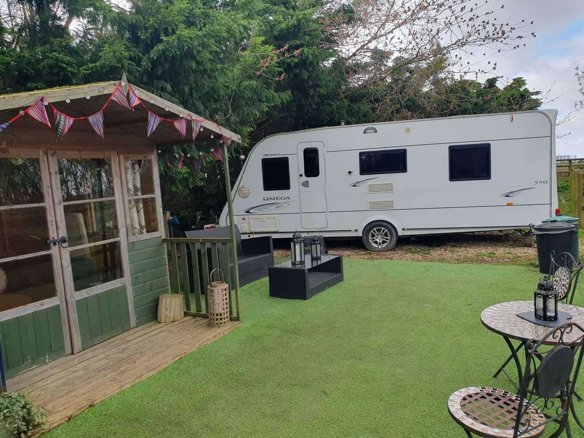 Una caravana estacionada en un patio trasero con una pequeña estructura de madera, una mesa de patio y césped.