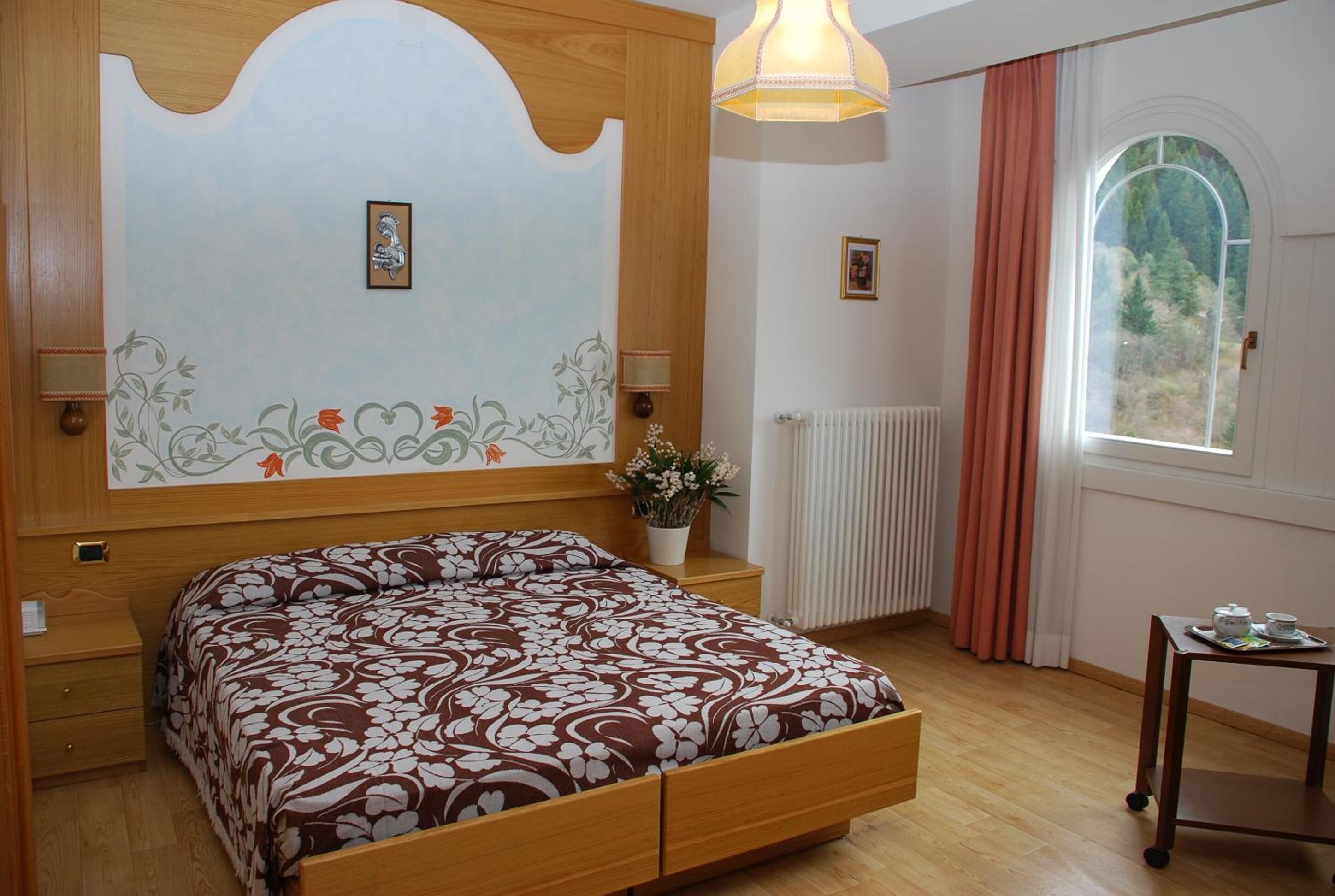 A bedroom with a wooden bed, a nightstand, a bedside table, a window with curtains, and a radiator.