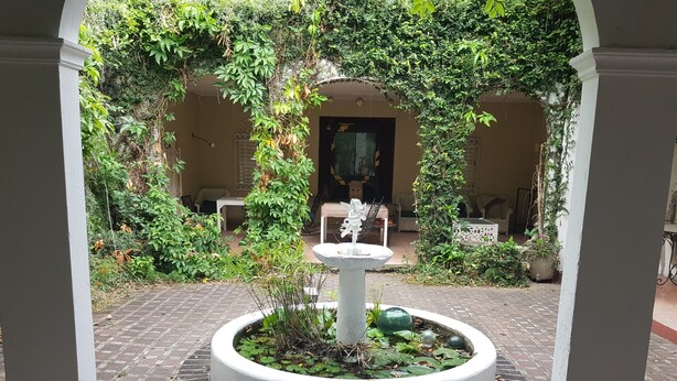 A courtyard with a central fountain surrounded by a circular brick border, under a covered area with arches and greenery.