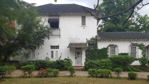 A two-story white building with a tiled roof, surrounded by greenery and a grassy area with scattered flowers.