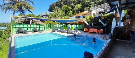 Une piscine entourée de verdure et de bâtiments, avec des chaises longues et des parasols.