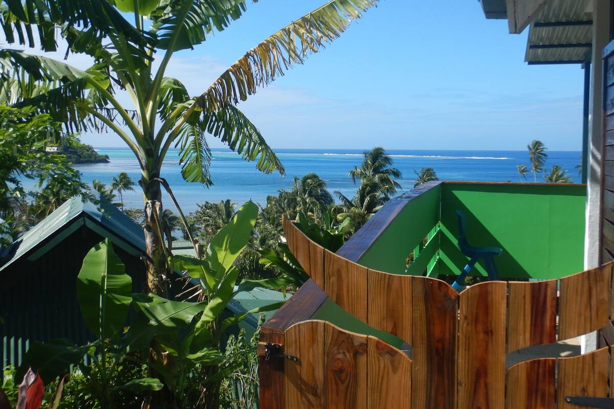 A wooden structure with a green interior, surrounded by tropical vegetation and overlooking a beach with palm trees and the ocean.