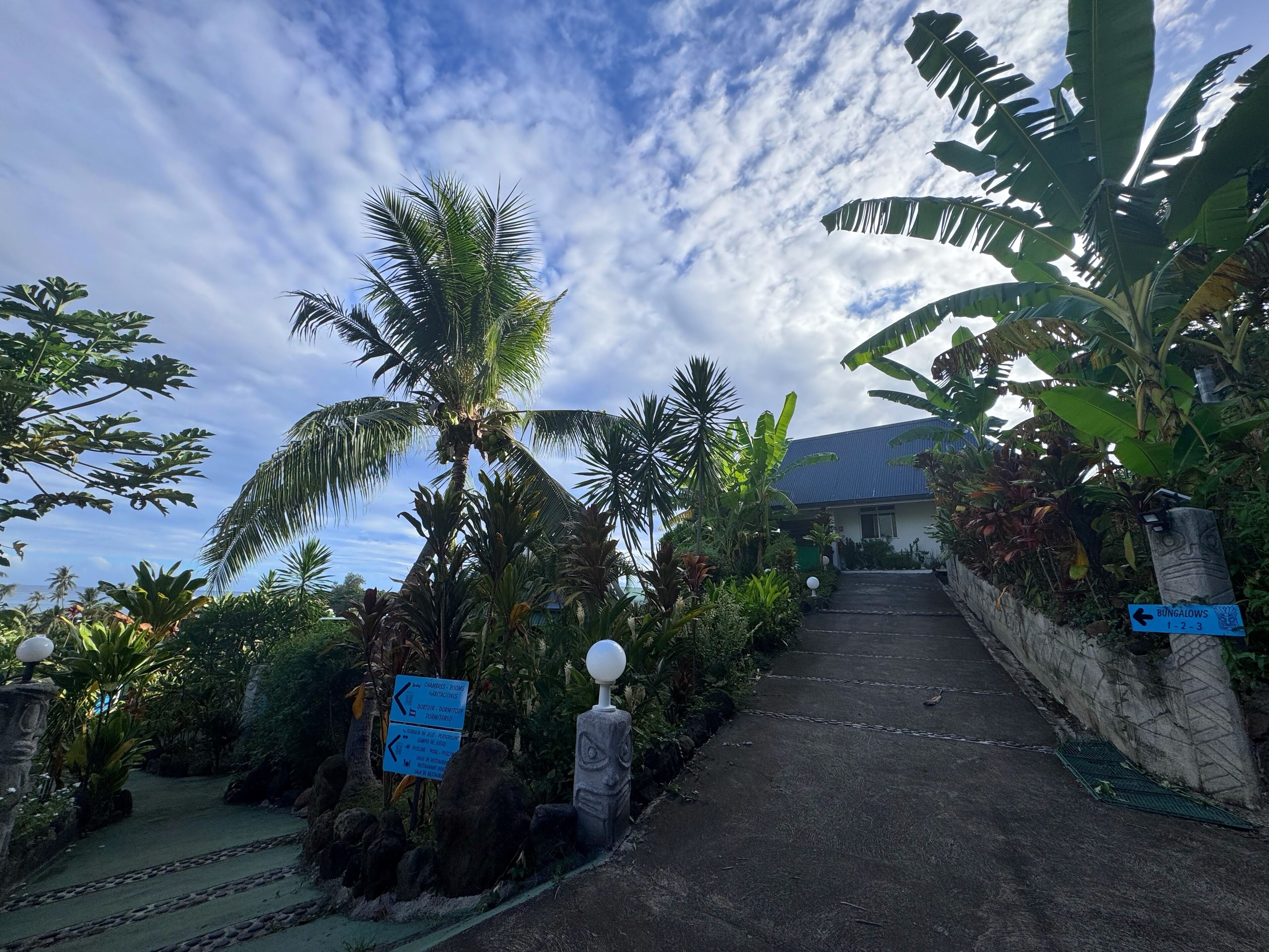 A pathway leading to a building surrounded by tropical vegetation, including palm trees and banana plants. There are directional signs indicating 'BUNGALOWS' and 'HOTEL'.