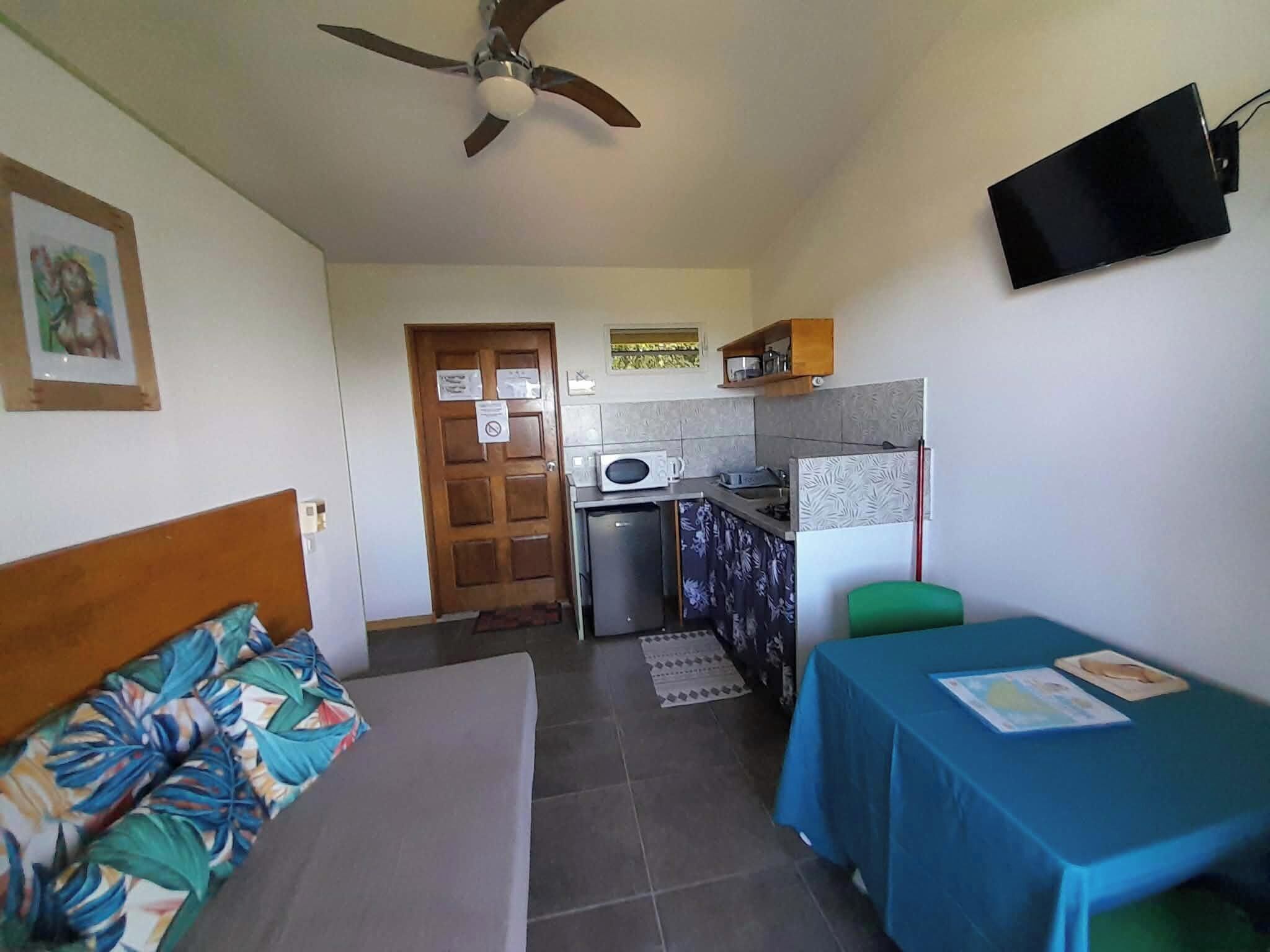 A compact room with a bed, a small table, a kitchenette, and a ceiling fan.