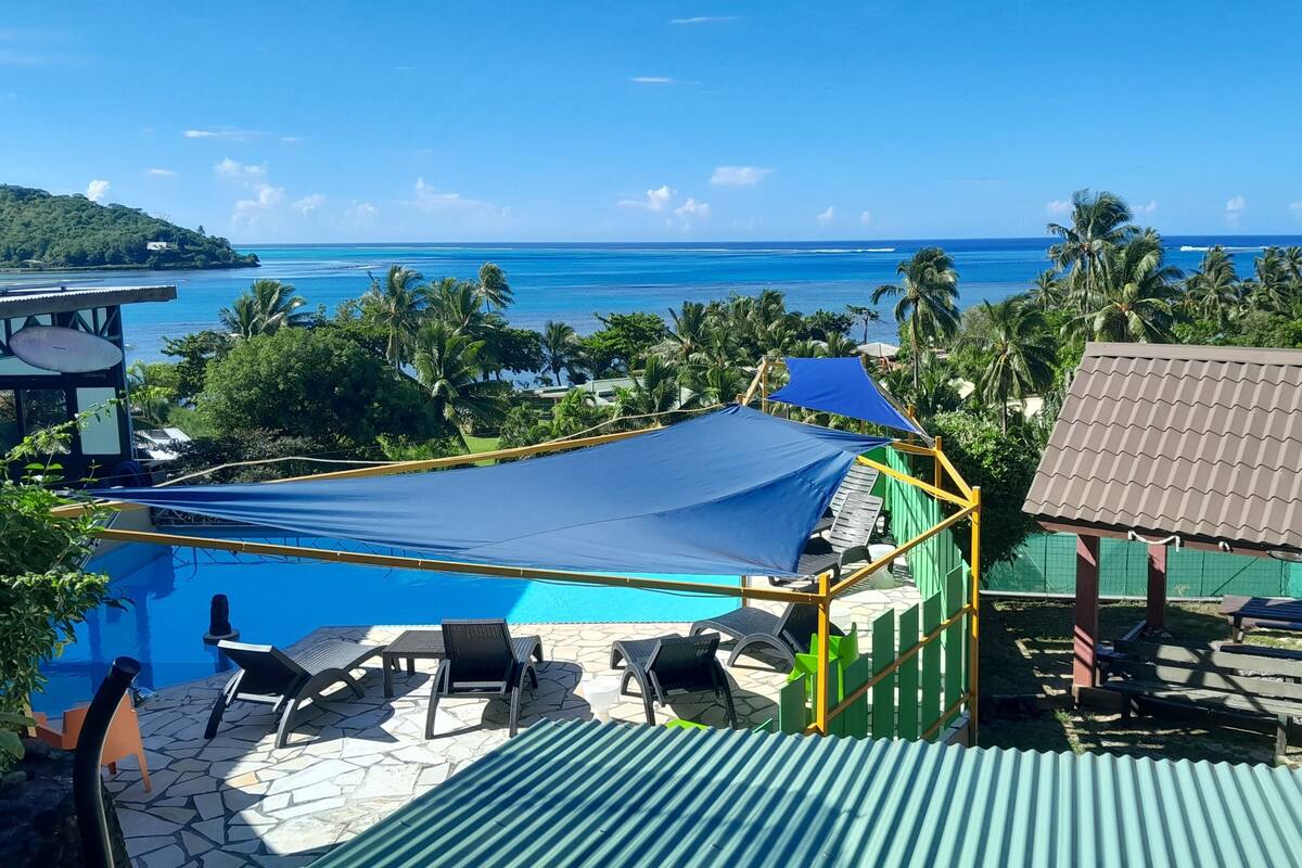 A pool area with lounge chairs and a shaded area, surrounded by tropical vegetation and a view of the ocean.