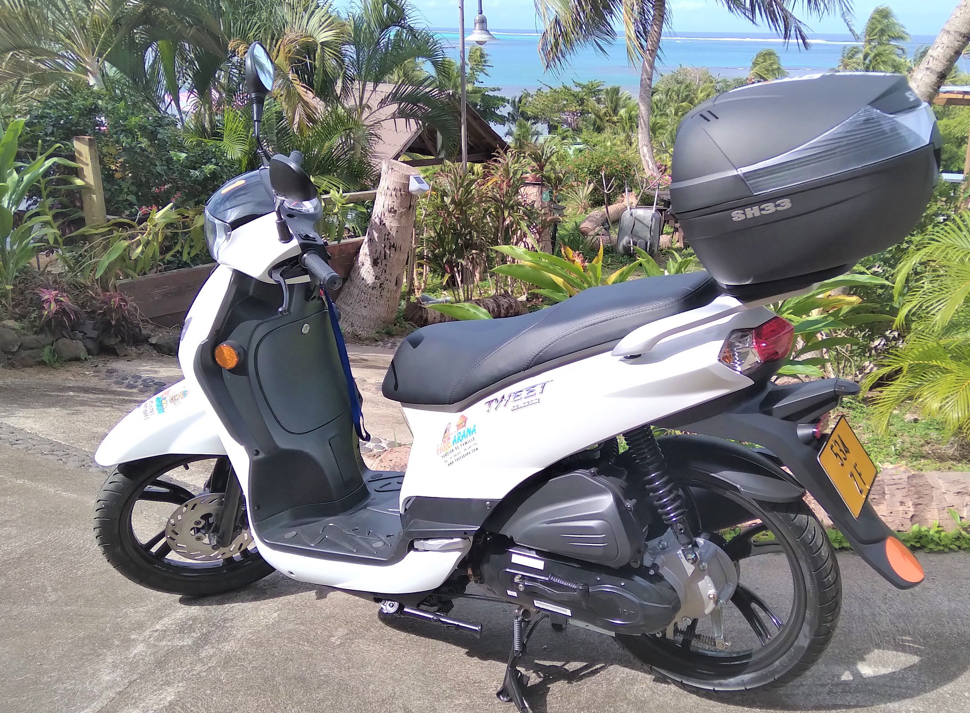 Un scooter blanc, garé à l’extérieur, avec une selle noire et un coffre de rangement à l’arrière.