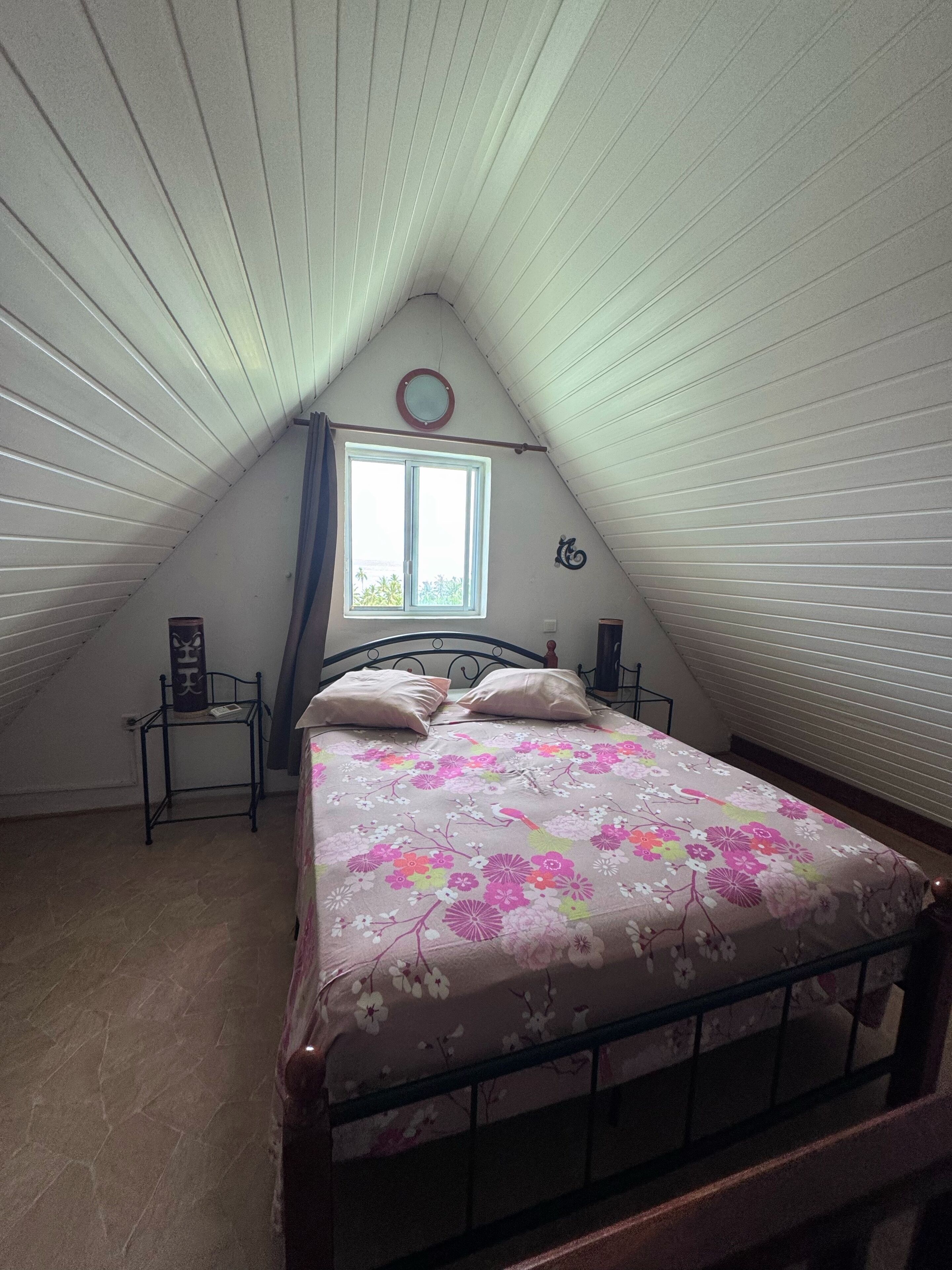 Um quarto com teto inclinado, uma cama com colcha florida, uma janela com vista e uma mesa pequena com um objeto decorativo.