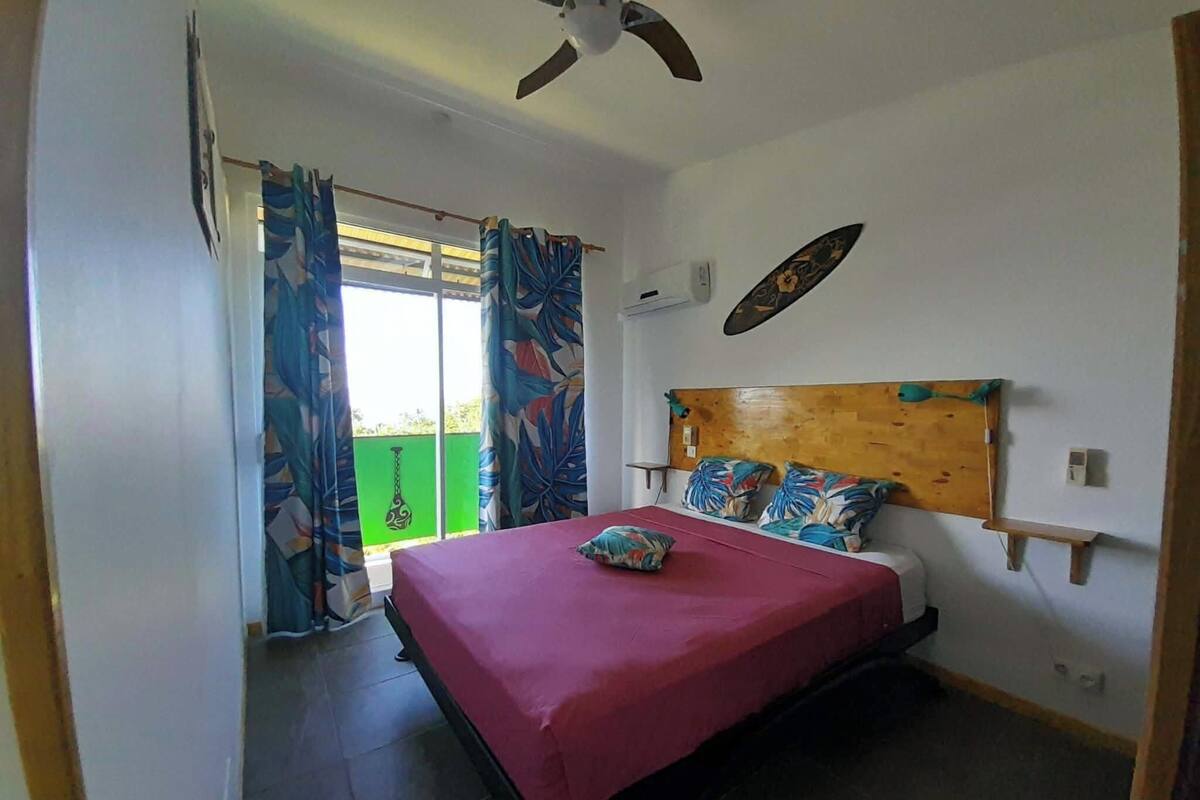 A bedroom with a bed, curtains, a ceiling fan, and a view of greenery outside.