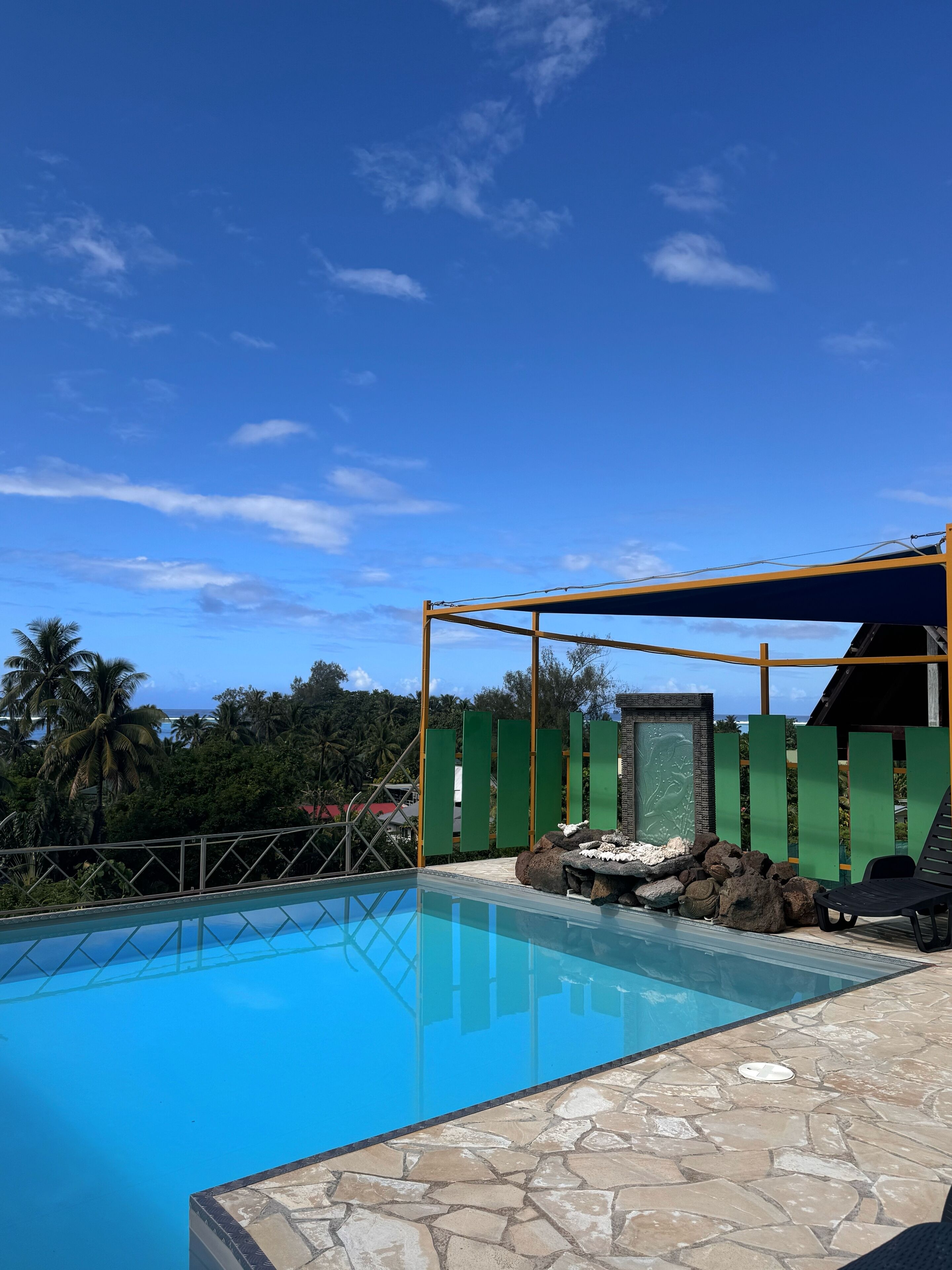 A clear blue pool with a stone-tiled deck, a covered seating area, and a view of the ocean and palm trees.