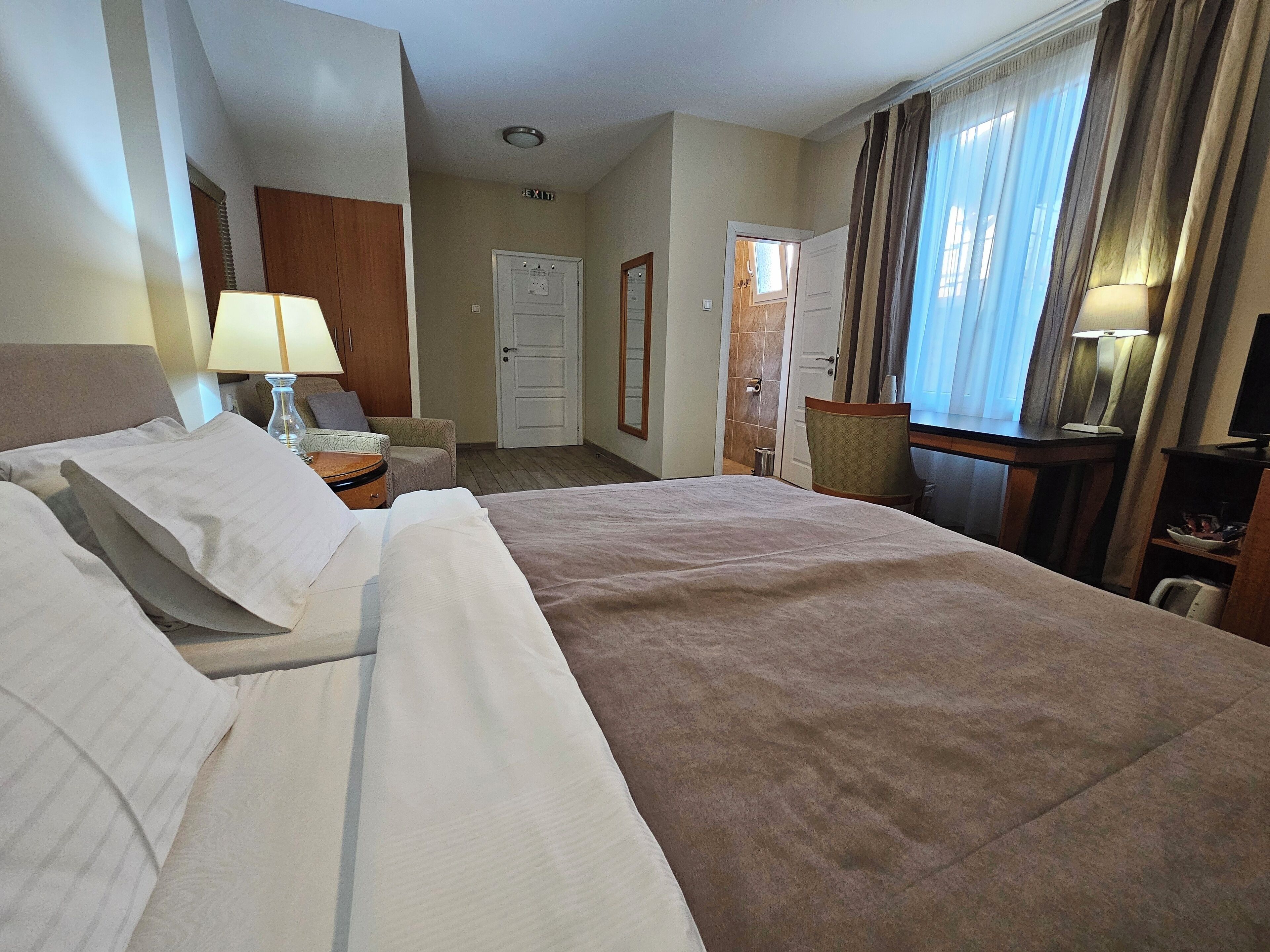 A hotel room with a large bed, bedside tables with lamps, a desk with a chair, a wardrobe, and a bathroom door.