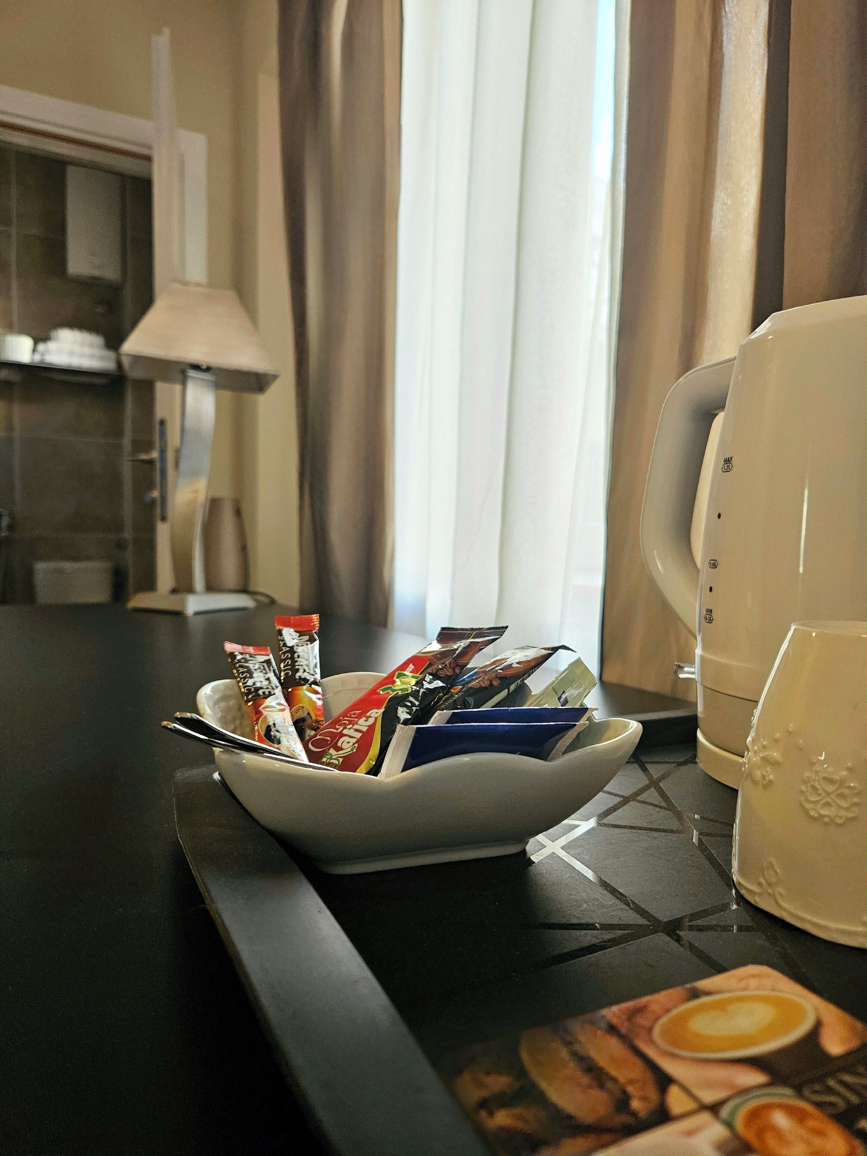 A hotel room with a bowl of snacks on the table, a coffee maker, and a window with curtains.