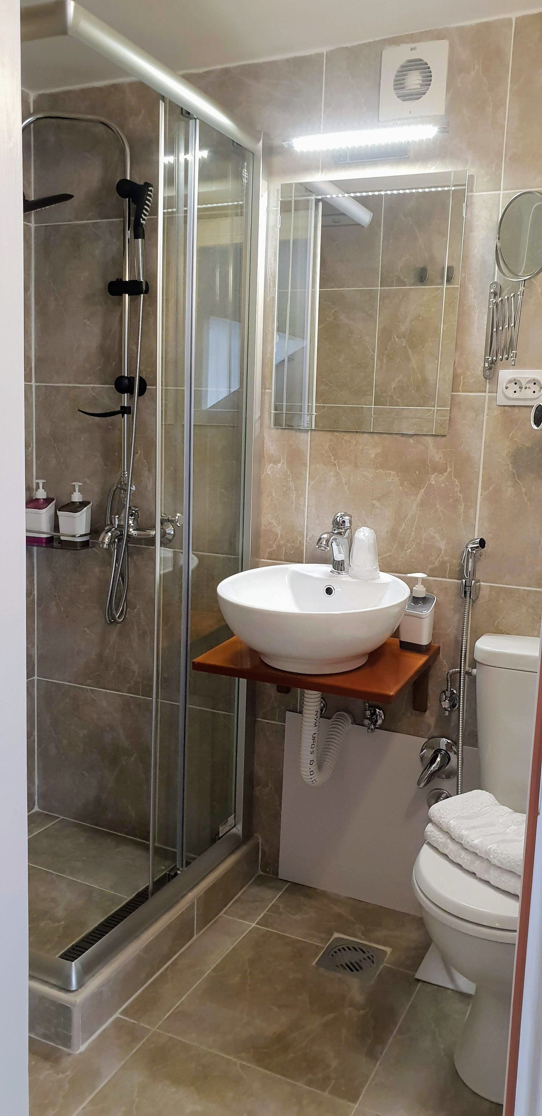 A modern bathroom with a glass shower enclosure, a white sink with a wooden shelf, a mirror, and a toilet.