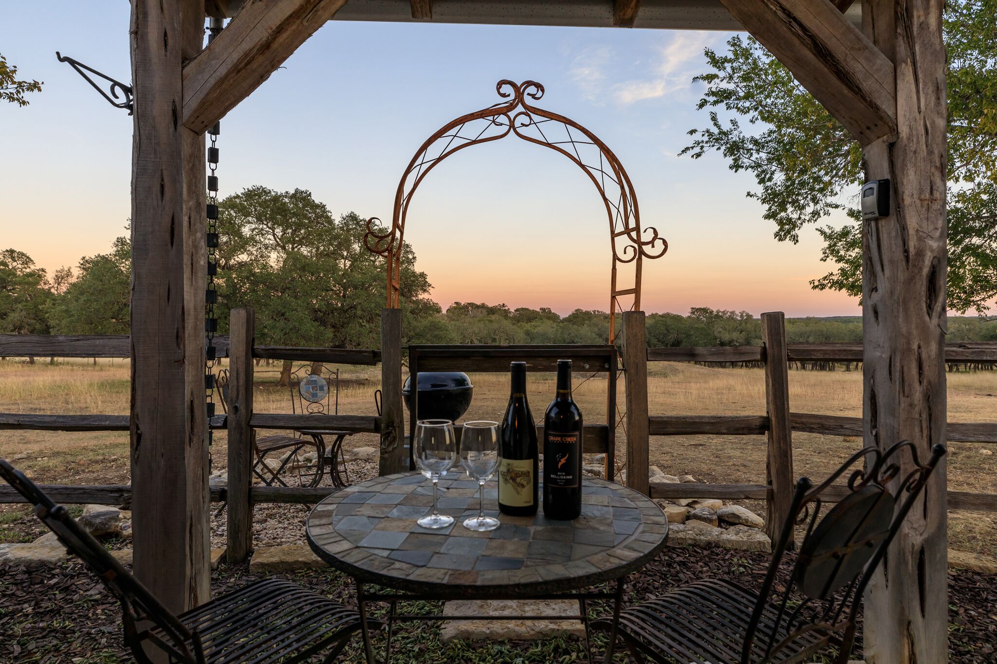 A rustic outdoor seating area with a metal table and chairs, a wooden archway, and a bottle of wine.